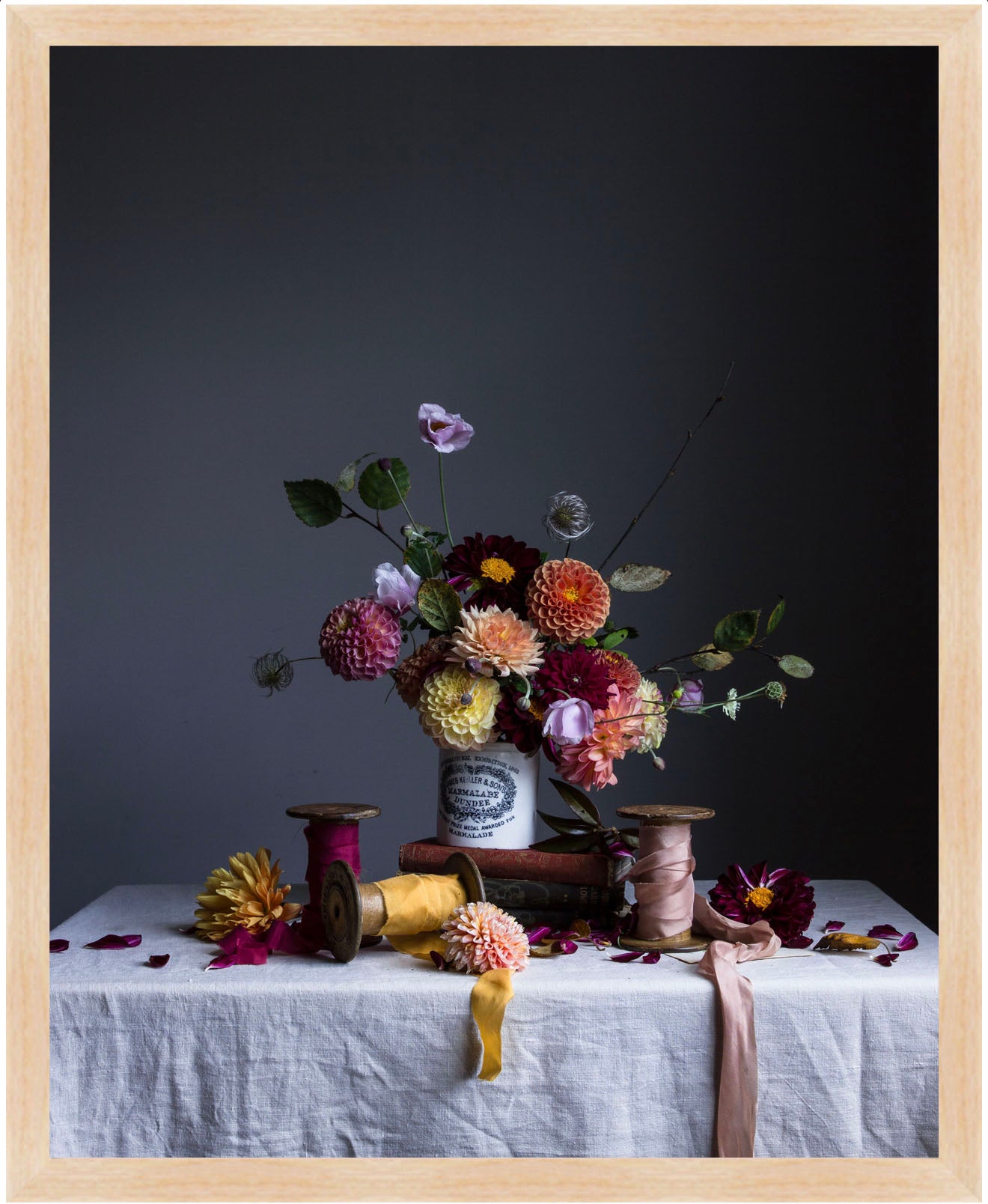 Still life arrangement with flowers, ribbons, and spools on a table against a dark background. Botanical art