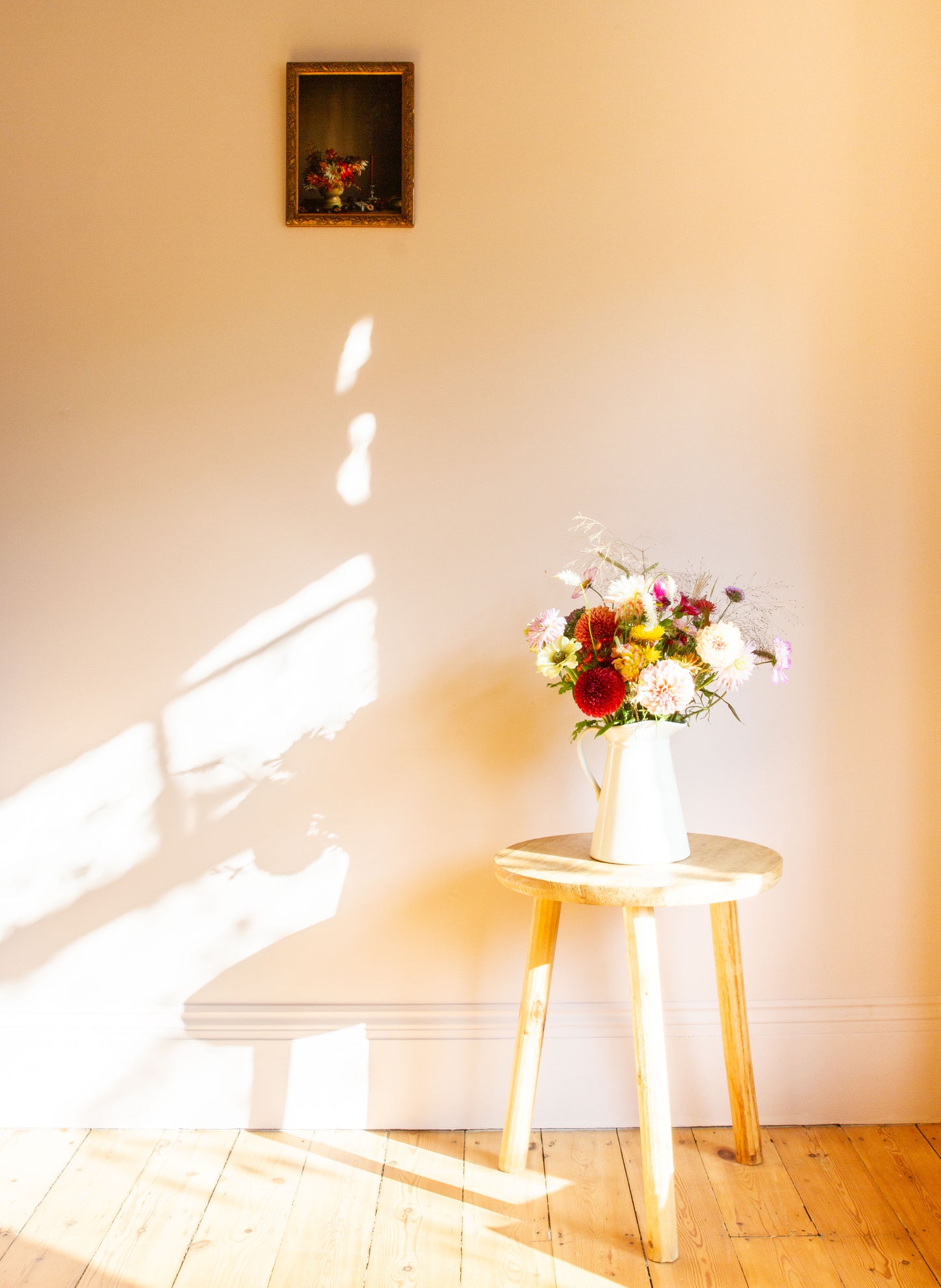 Floral arrangement on a small table with a light source casting shadows on a wall alongside a framed limited edition floral still-life by Emma Harris