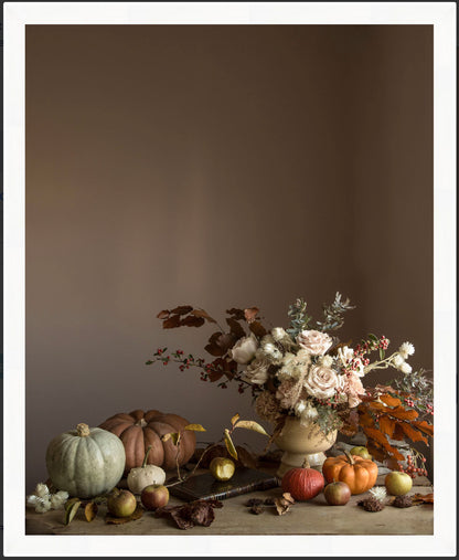 Framed botanical art with pumpkins, apples, and a floral bouquet on a wooden surface against a brown background.