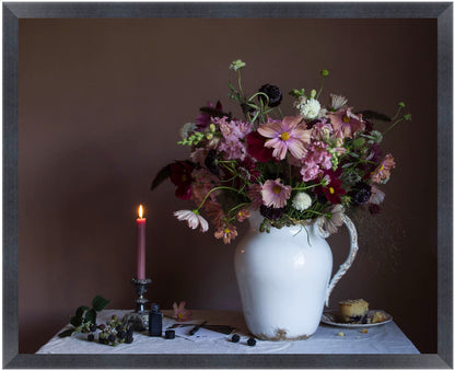 Framed still life arrangement with flowers in a white vase, candle, and small plate on a table against a dark background.
