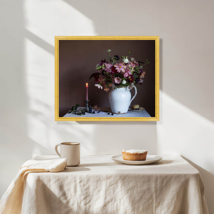 Framed artwork of a floral arrangement on a wall above a table with a pie and mug.