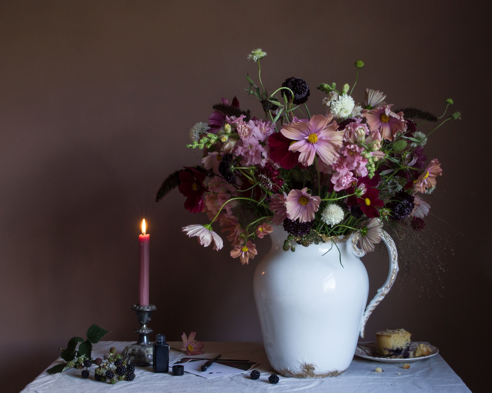Still-life fine art photography by Emma Harris showing a floral arrangement in a white vase with a lit candle and small plate on a table against a dark background