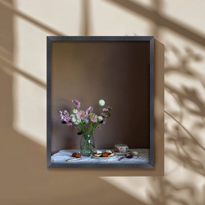 Framed still life with flowers and food on a table against a neutral wall.