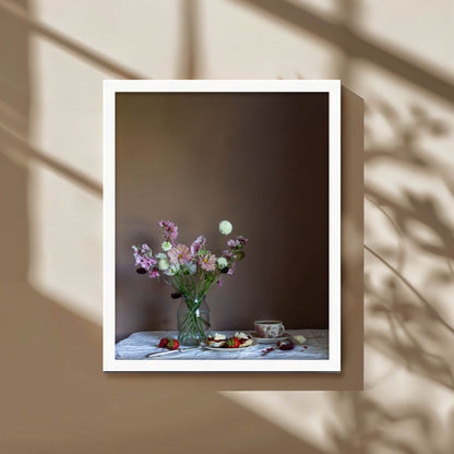 Framed photograph of a still life scene with flowers and food on a table against a brown background.