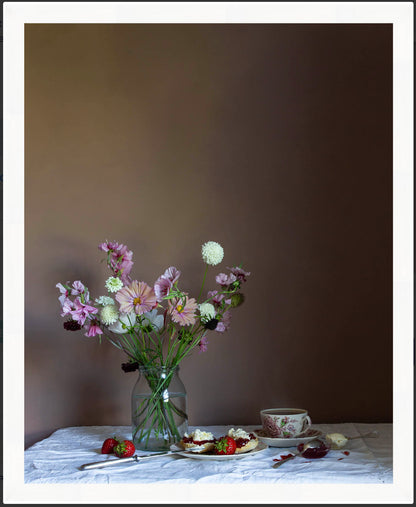 Framed still life with flowers in a vase, a teacup, and dessert on a table against a brown wall.
