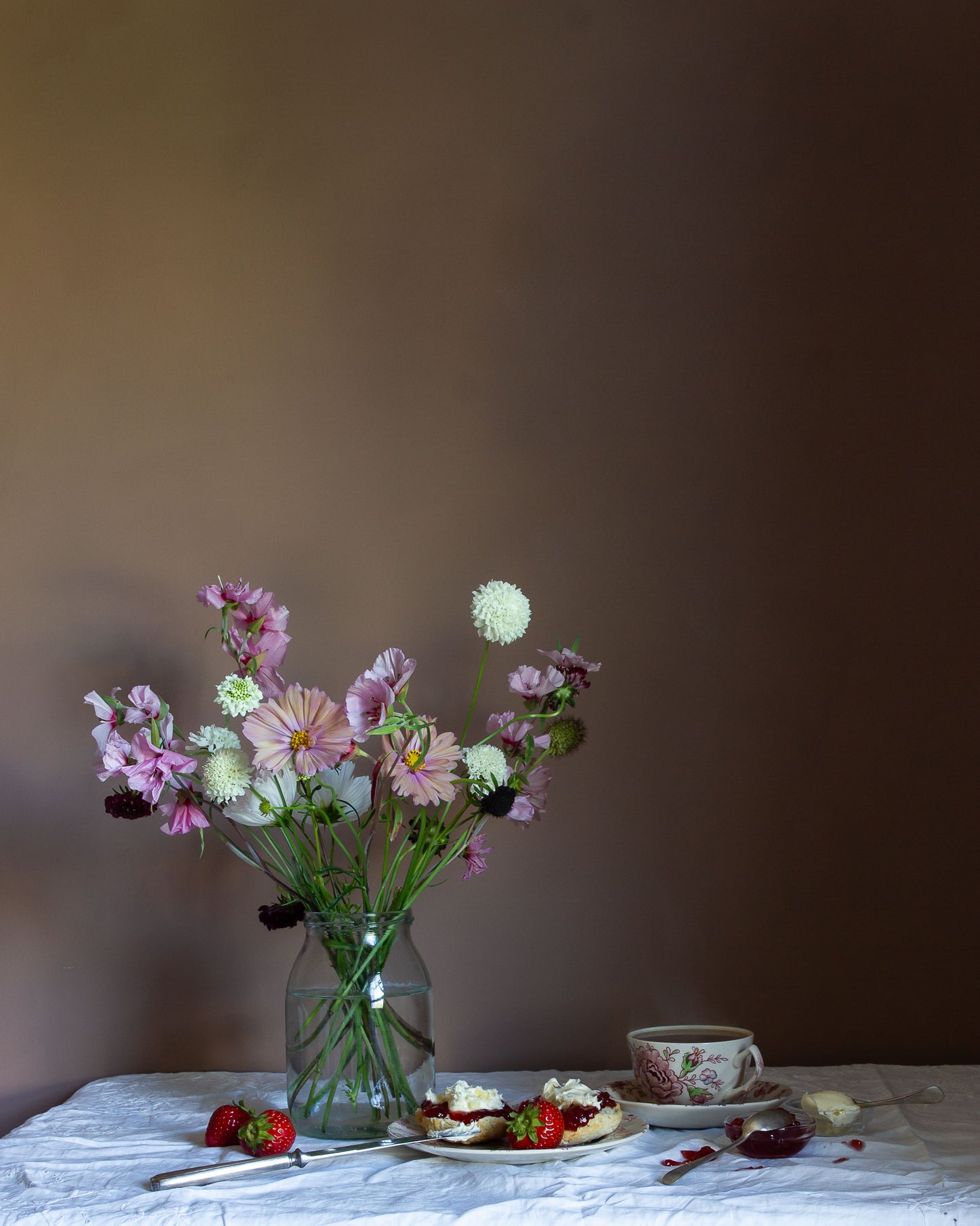 Still-life photograph of a bouquet of flowers in a glass vase on a table with a cake and strawberries against a plain background by fine art photographer Emma Harris