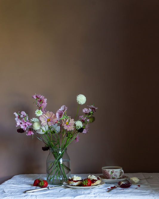 Still-life photograph of a bouquet of flowers in a glass vase on a table with a cake and strawberries against a plain background by fine art photographer Emma Harris