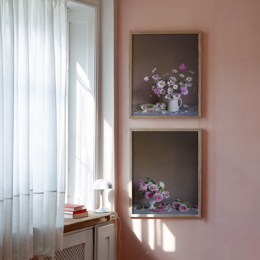 Two framed still life floral art prints by photographer Emma Harris with pink and white floral arrangements hang on a dusty pink wall next to a sunlit window with white curtains.
