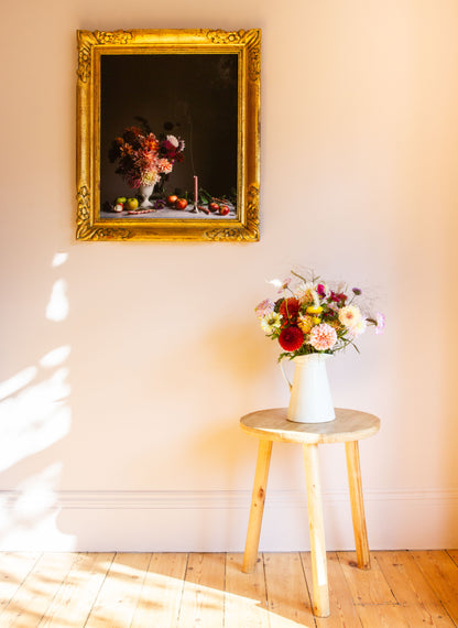 Decorative still life floral photograph on a wall with a vase of flowers on a stool below.