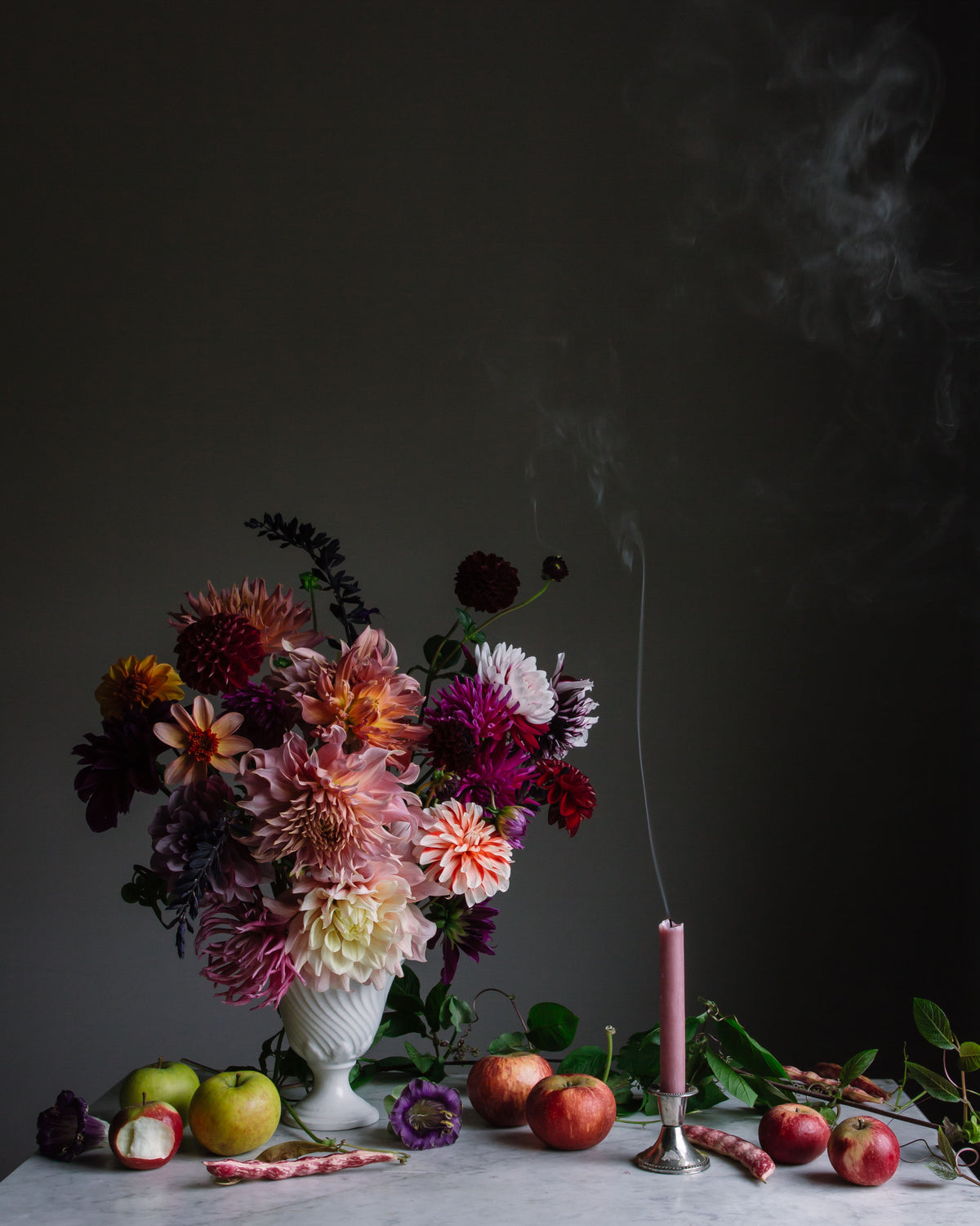 A fine art floral photograph featuring a still life composition with a vibrant bouquet of dahlias in a white vase, accompanied by beans, apples, and a smoking candle, set against a dark background by Emma Harris