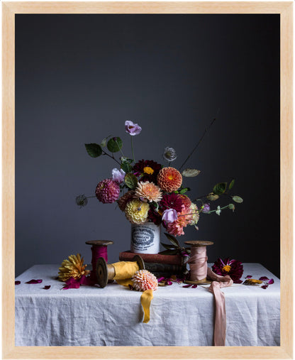 Still life arrangement with flowers, ribbons, and spools on a table against a dark background.   Botanical art