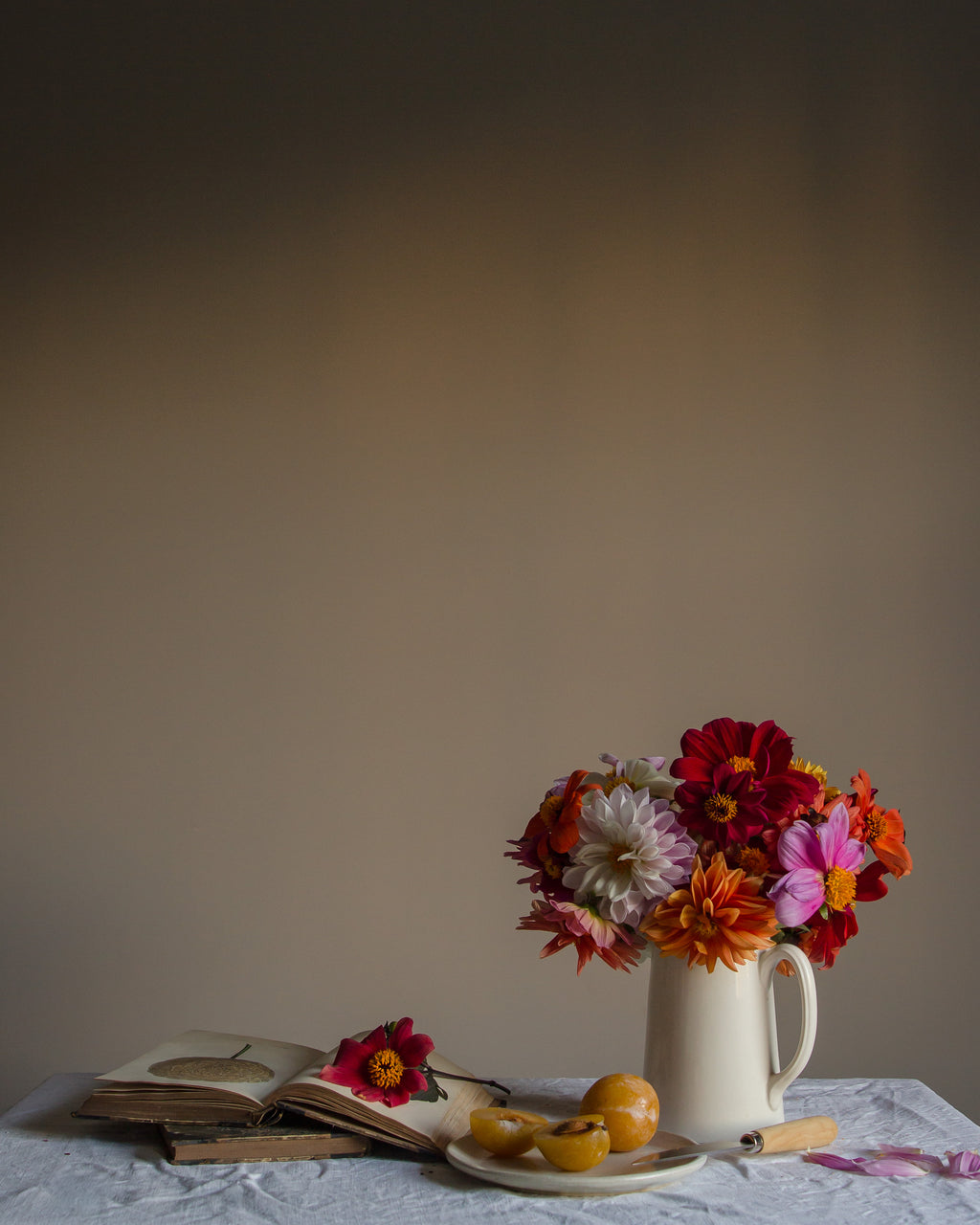 A limited edition fine art photographic print featuring a bouquet of dahlias and plums in a ceramic jug, with a book and a few scattered petals on a table, against a neutral background by Emma Harris