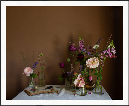 Framed photographic still life image showing an arrangement of flowers in vases on a table with a brown background