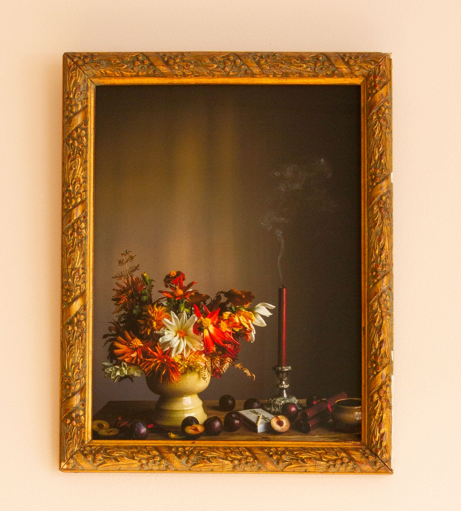 Decorative still life scene with flowers and candle in a gold frame on a beige wall.