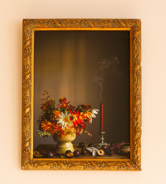 Decorative still life scene with flowers and candle in a gold frame on a beige wall.
