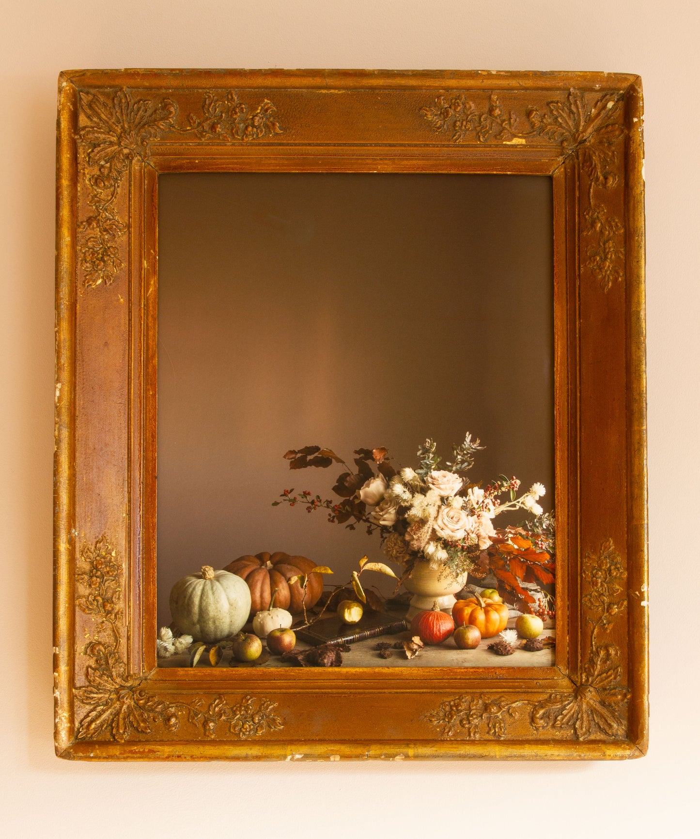 Decorative still life photograph with flowers and fruits in a gold frame on a beige wall.