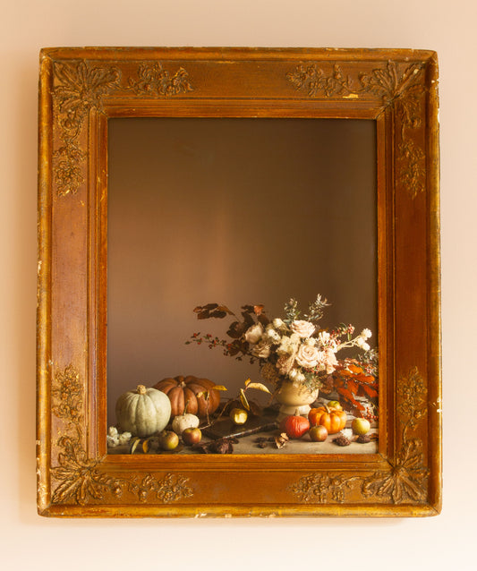 Decorative still life photograph with flowers and fruits in a gold frame on a beige wall.