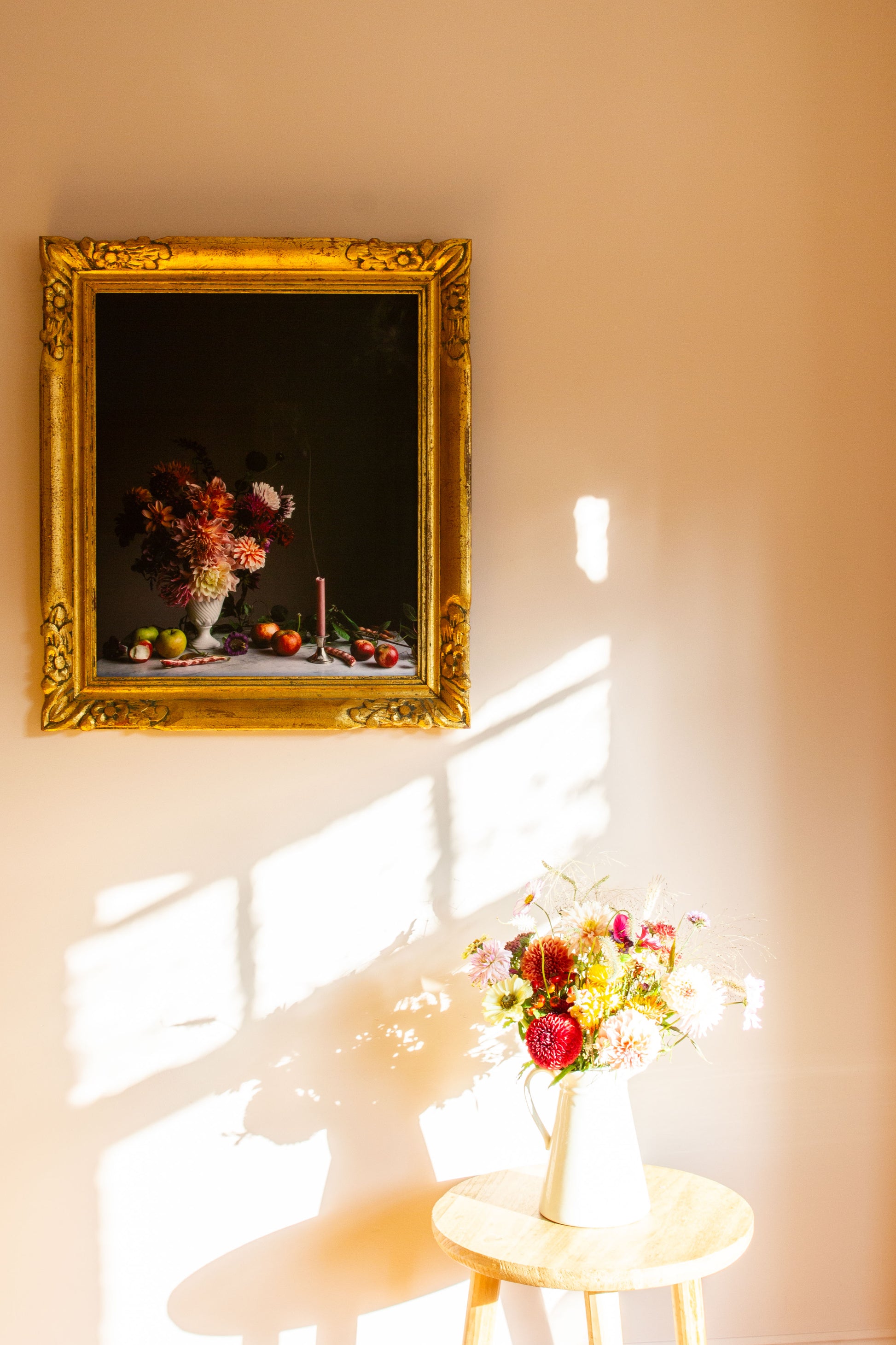 Gold-framed still-life floral photograph on a wall with a vase of flowers on a table below.