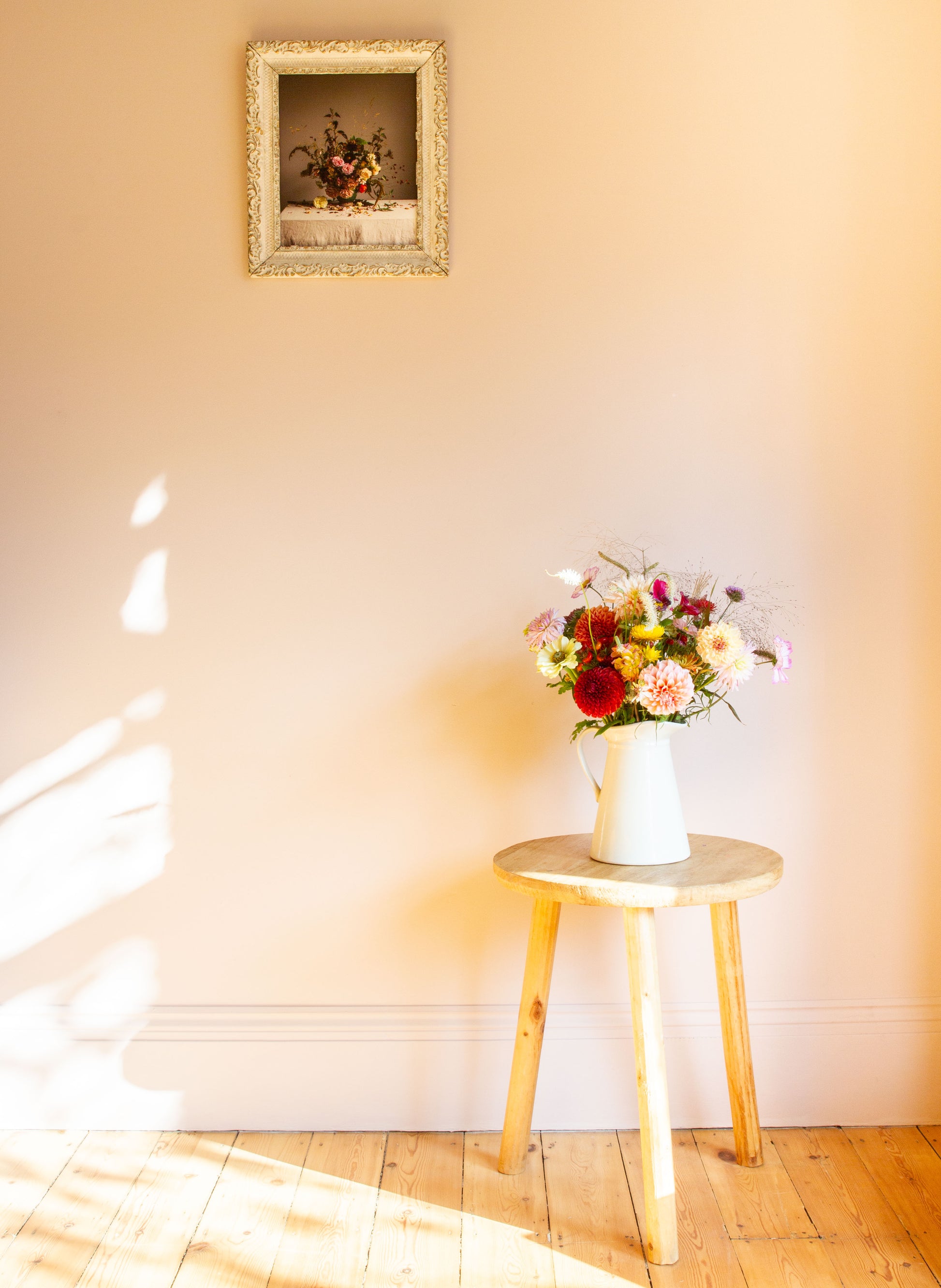 Floral arrangement in a white vase on a wooden stool against a light wall with a framed floral limited edition photograph.