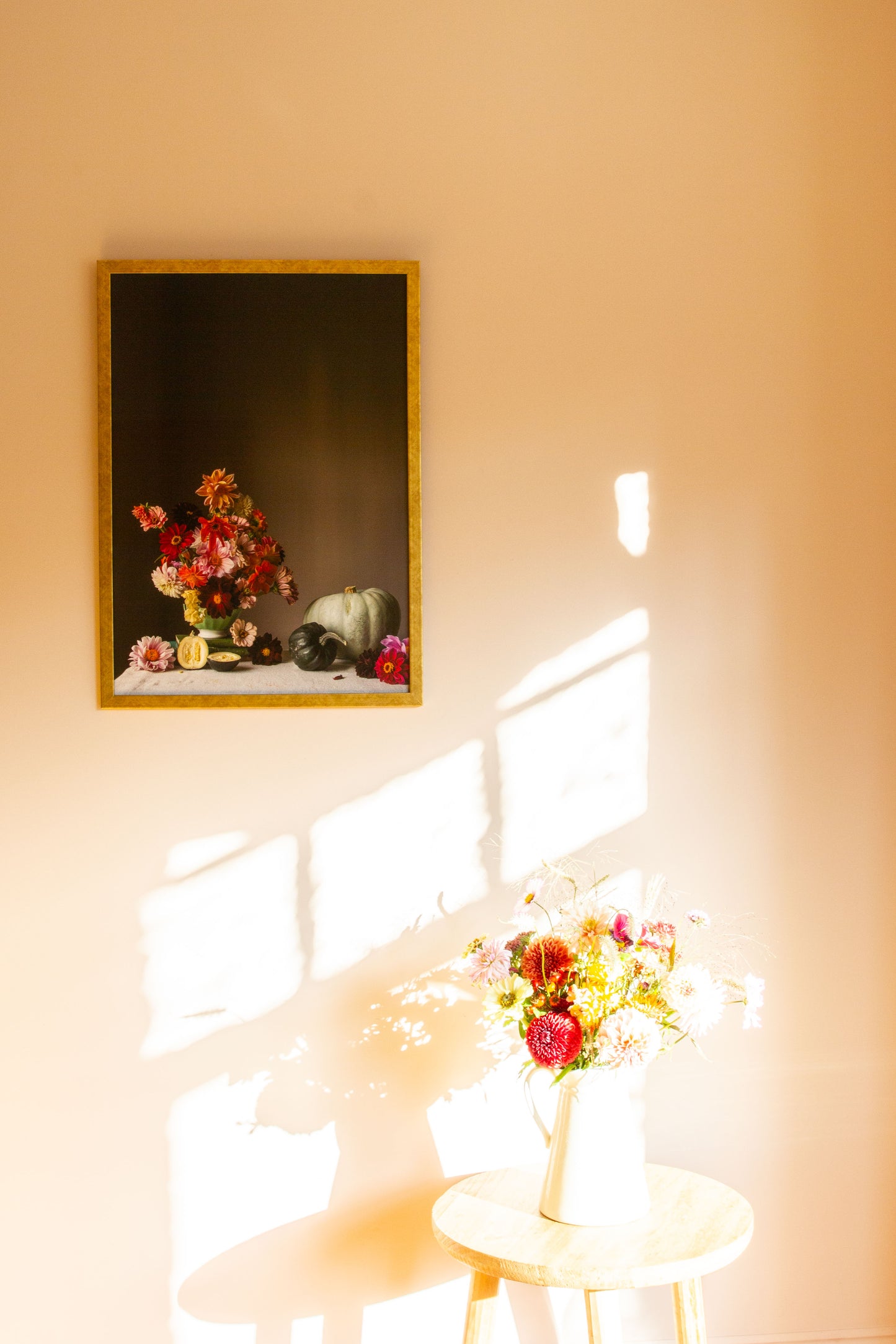 Framed artwork of flowers on a wall with a smaller vase of flowers on a table below.