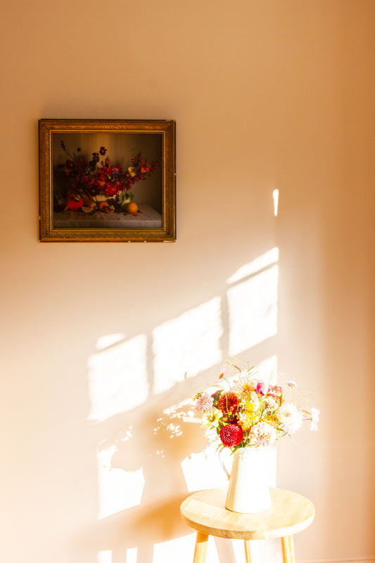 Framed floral artwork on a wall with a vase of flowers on a stool below.
