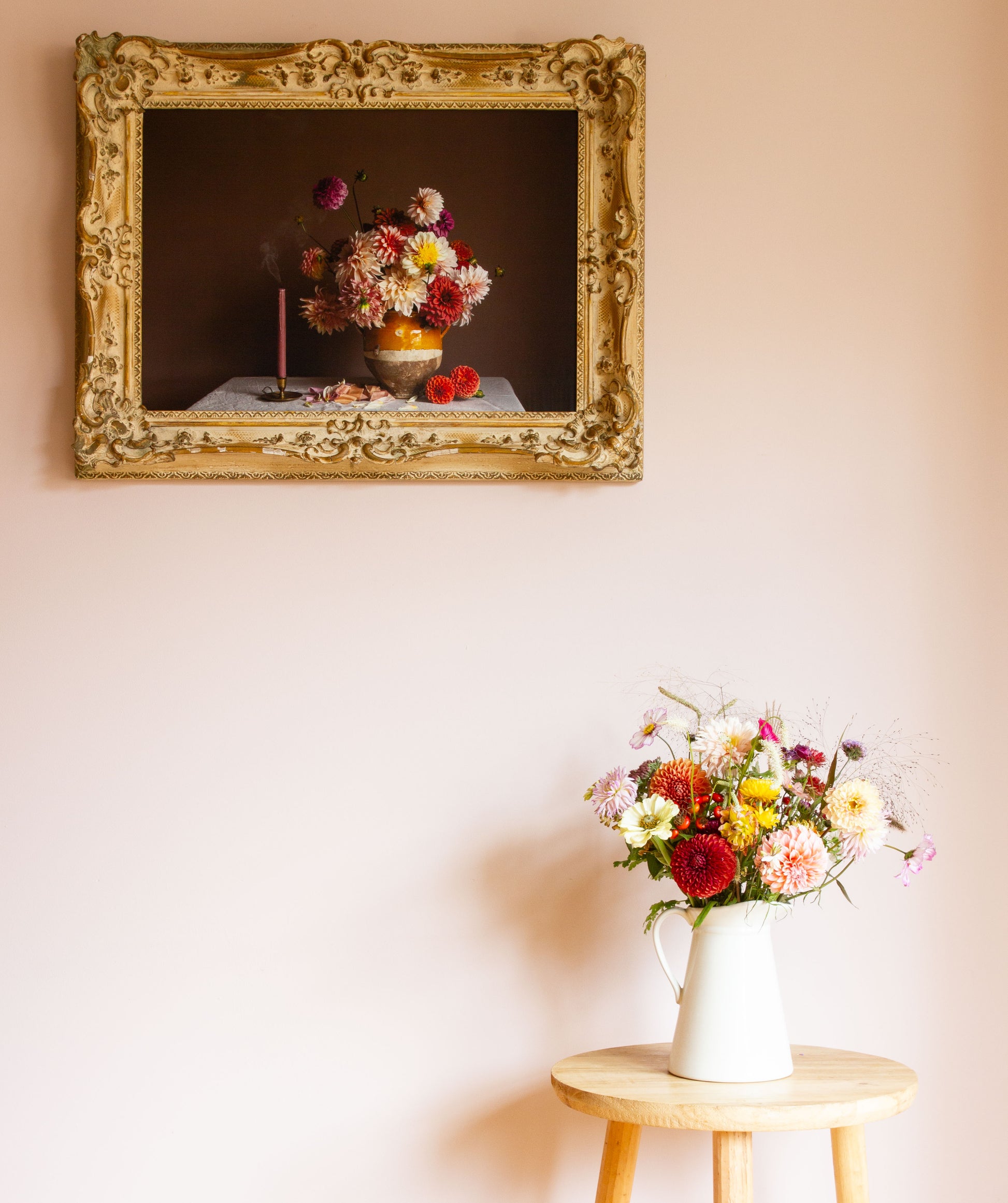 Floral arrangement in a white vase on a wooden table with a framed still-life photo of dahlias in the background.