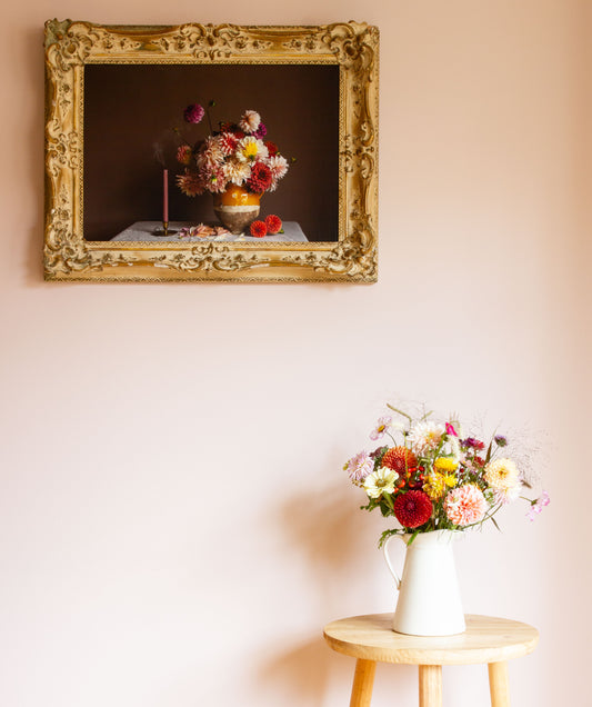 Floral arrangement in a white vase on a wooden table with a framed still-life photo of dahlias in the background.
