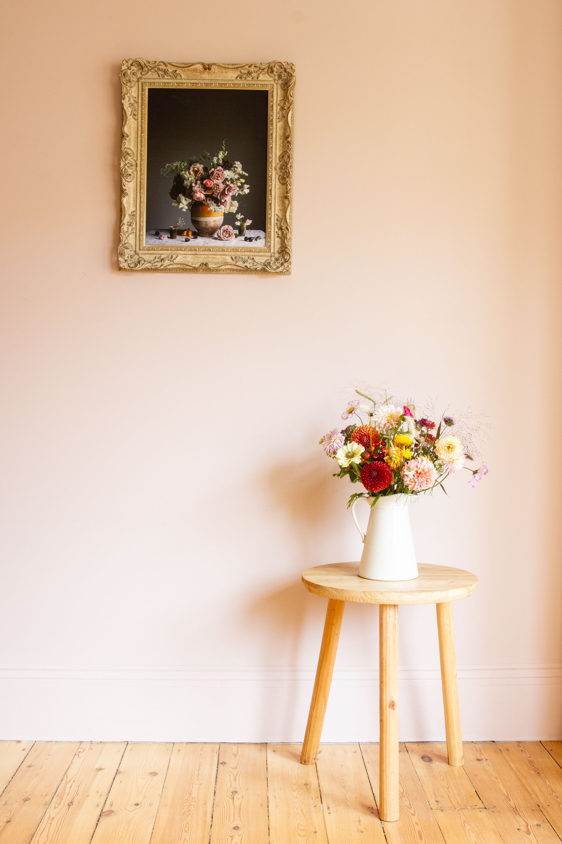 Floral arrangement on a small wooden table with a framed picture of still-life of flowers on the wall.