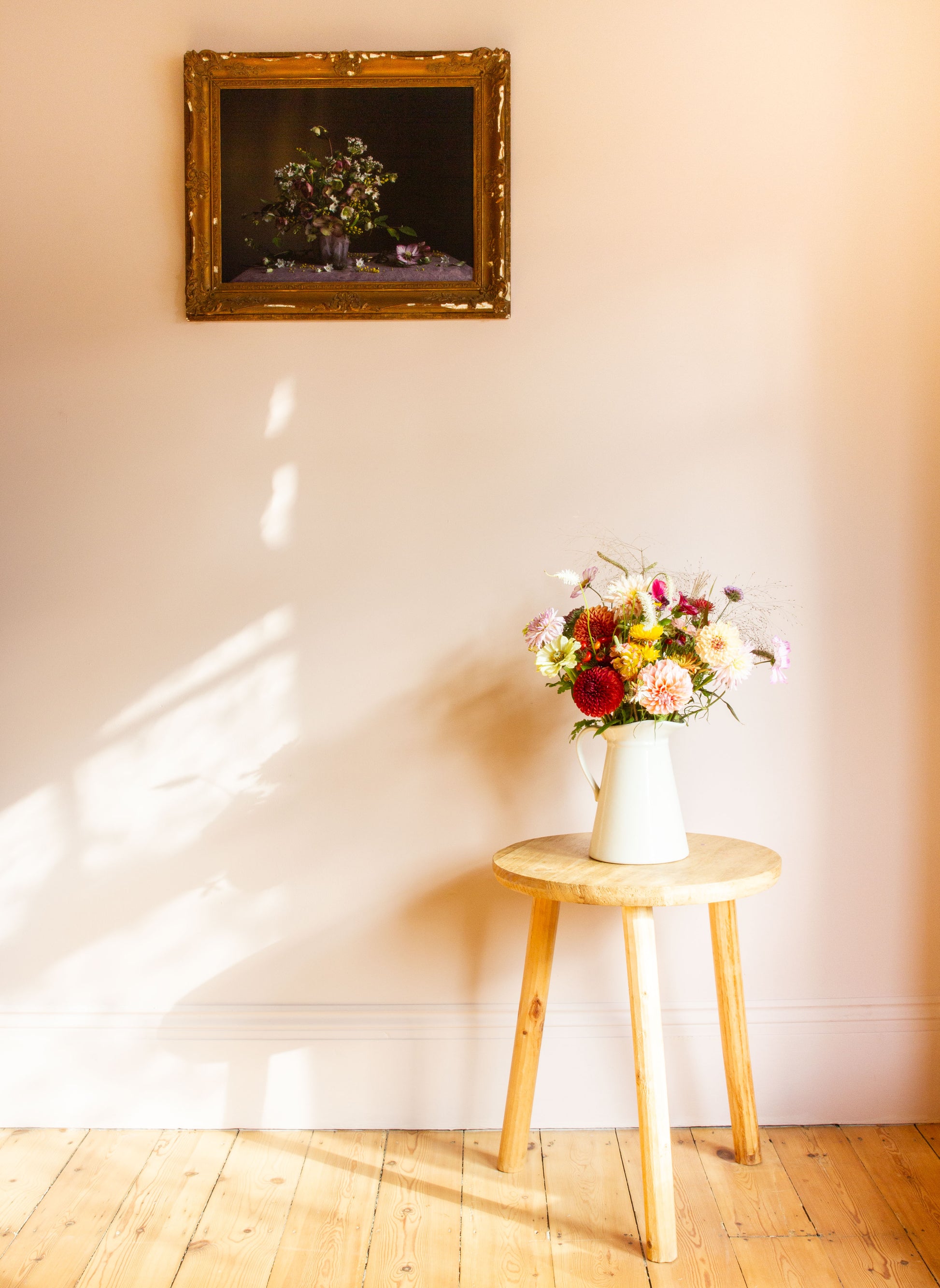 Floral arrangement in a white vase on a wooden stool against a wall with a framed picture.