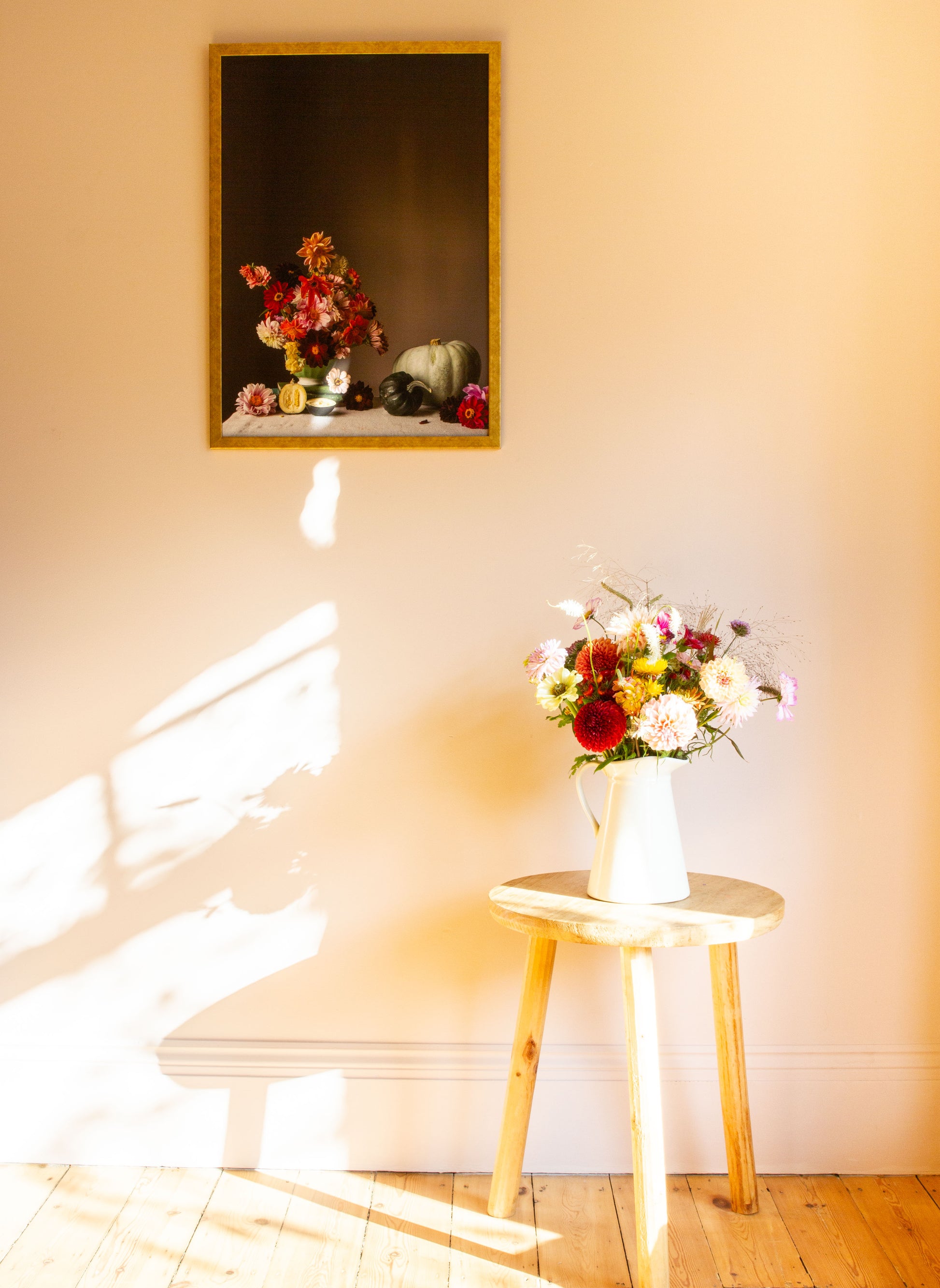 Floral arrangement in a vase on a small wooden table with a framed still-life photograph of flowers on the wall.