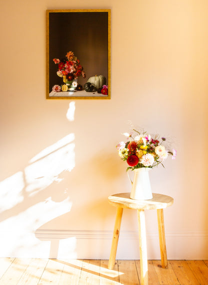 Floral arrangement in a vase on a small wooden table with a framed still-life photograph of flowers on the wall.