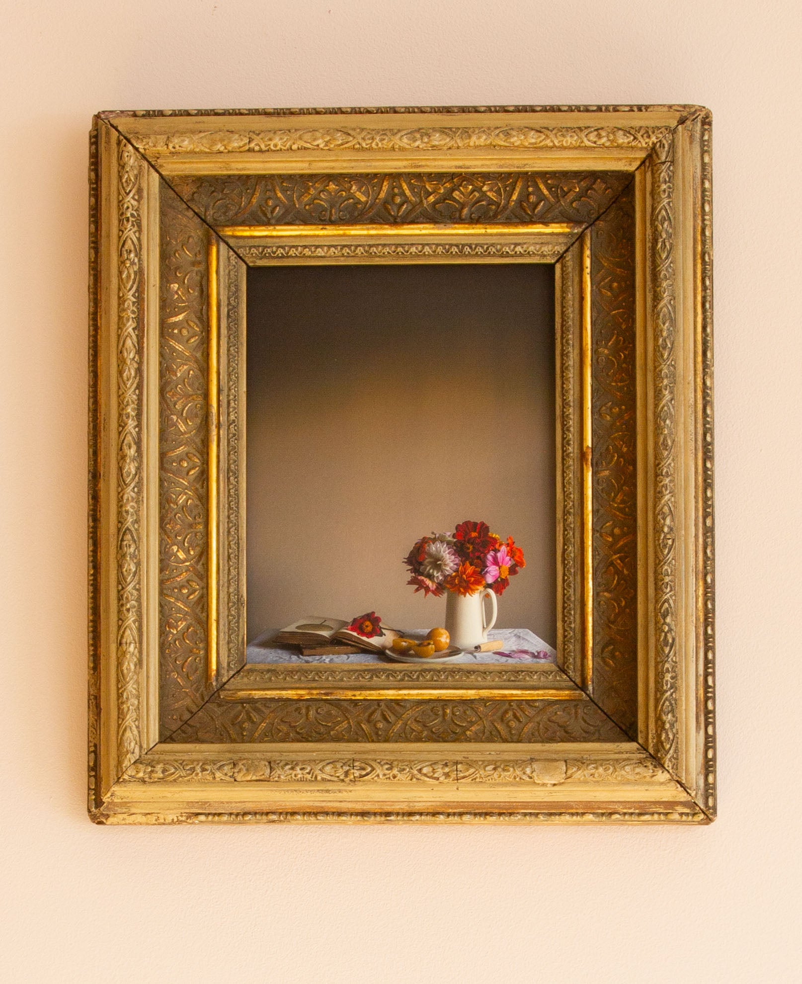 Decorative still life scene with flowers and fruit in a gold frame on a beige wall.
