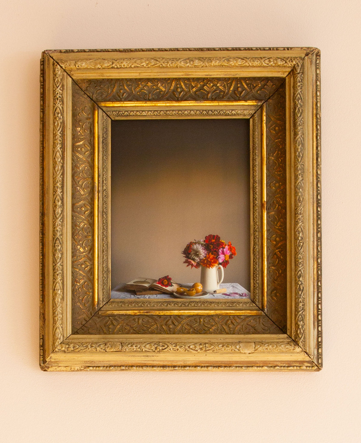 Decorative still life scene with flowers and fruit in a gold frame on a beige wall.