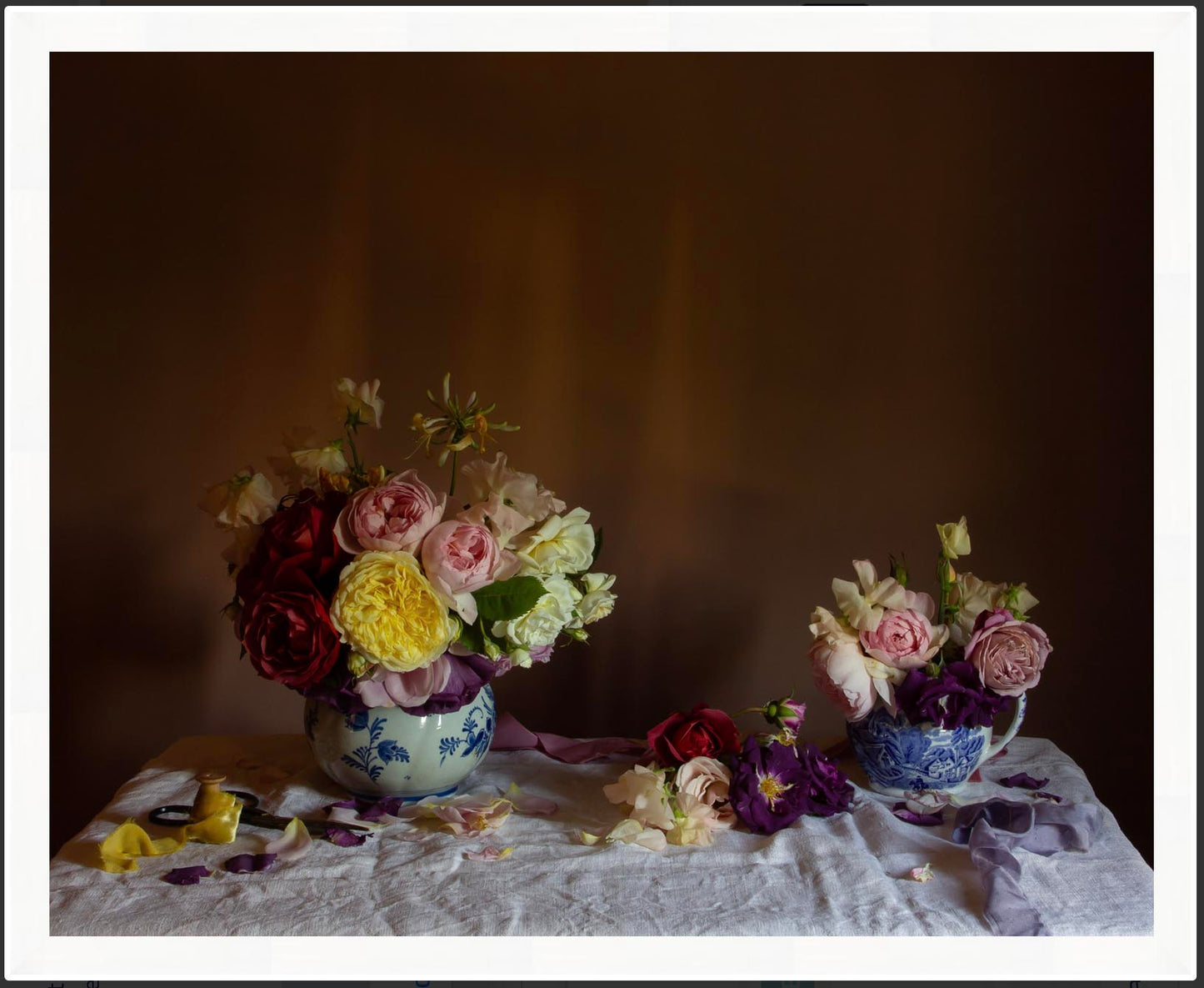 Framed floral still-life of two flower arrangement using roses in blue vases on a textured surface with a dark background in a white frame.