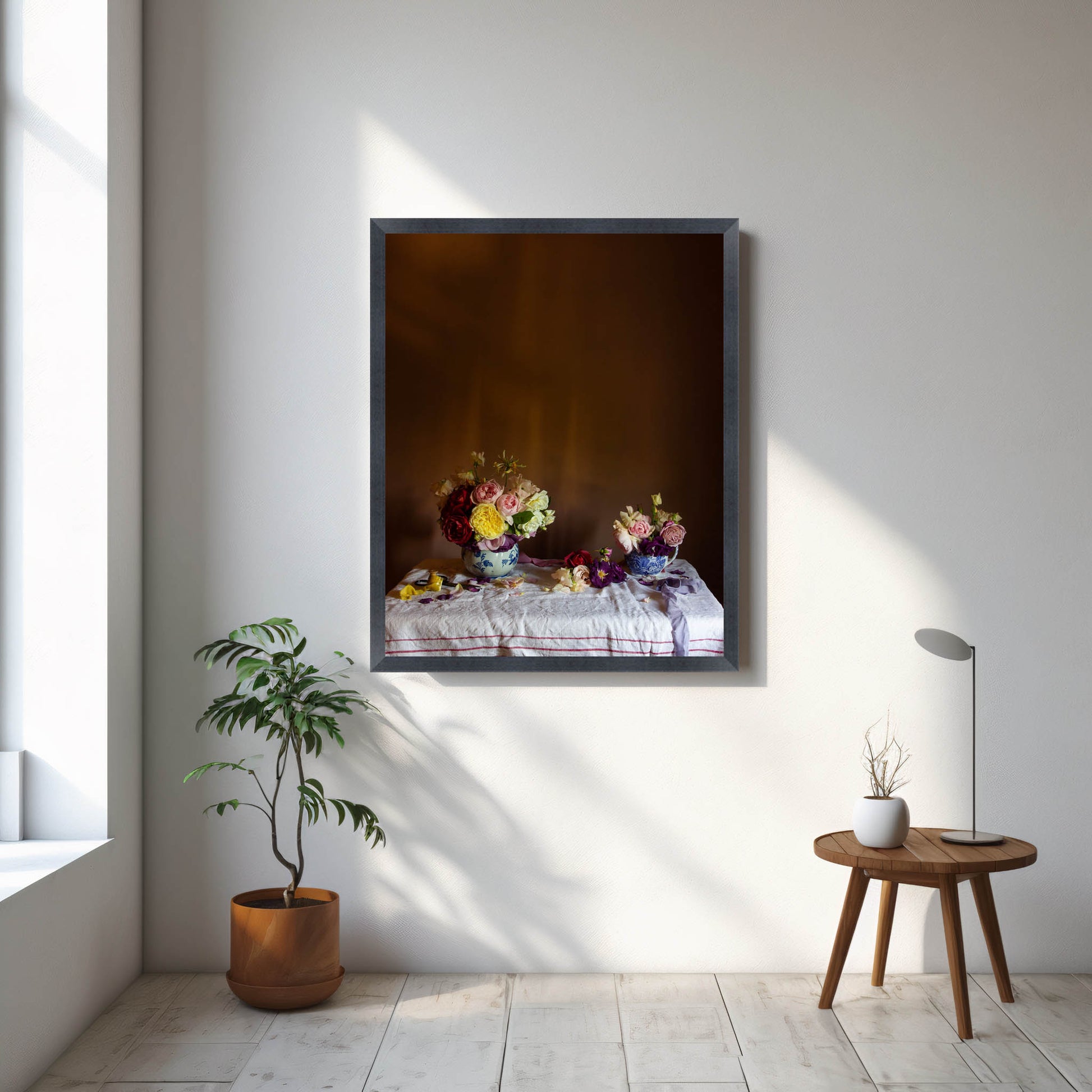 Framed artwork of flowers on a table in a room with a plant and small table.