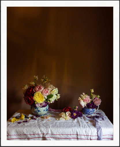 Framed still-life of floral arrangements in blue and white vases on a tablecloth against a brown background