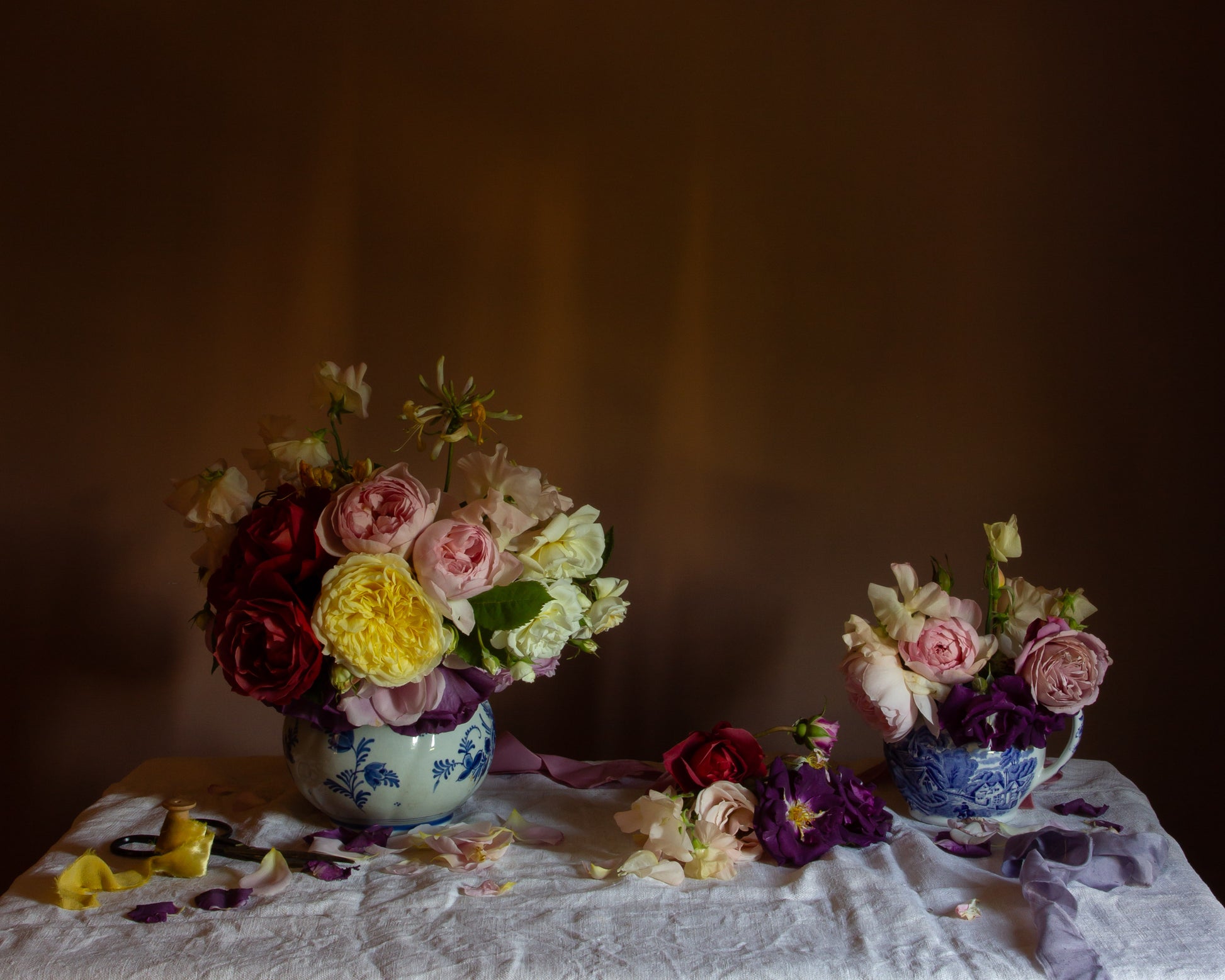 Still life floral with roses in blue and white jugs, on a white table cloth.