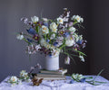 A fine art floral photograph by Emma Harris featuring a bouquet of various flowers including fritillary and grape hyacinths, displayed in a white vase, with books and a dark background.