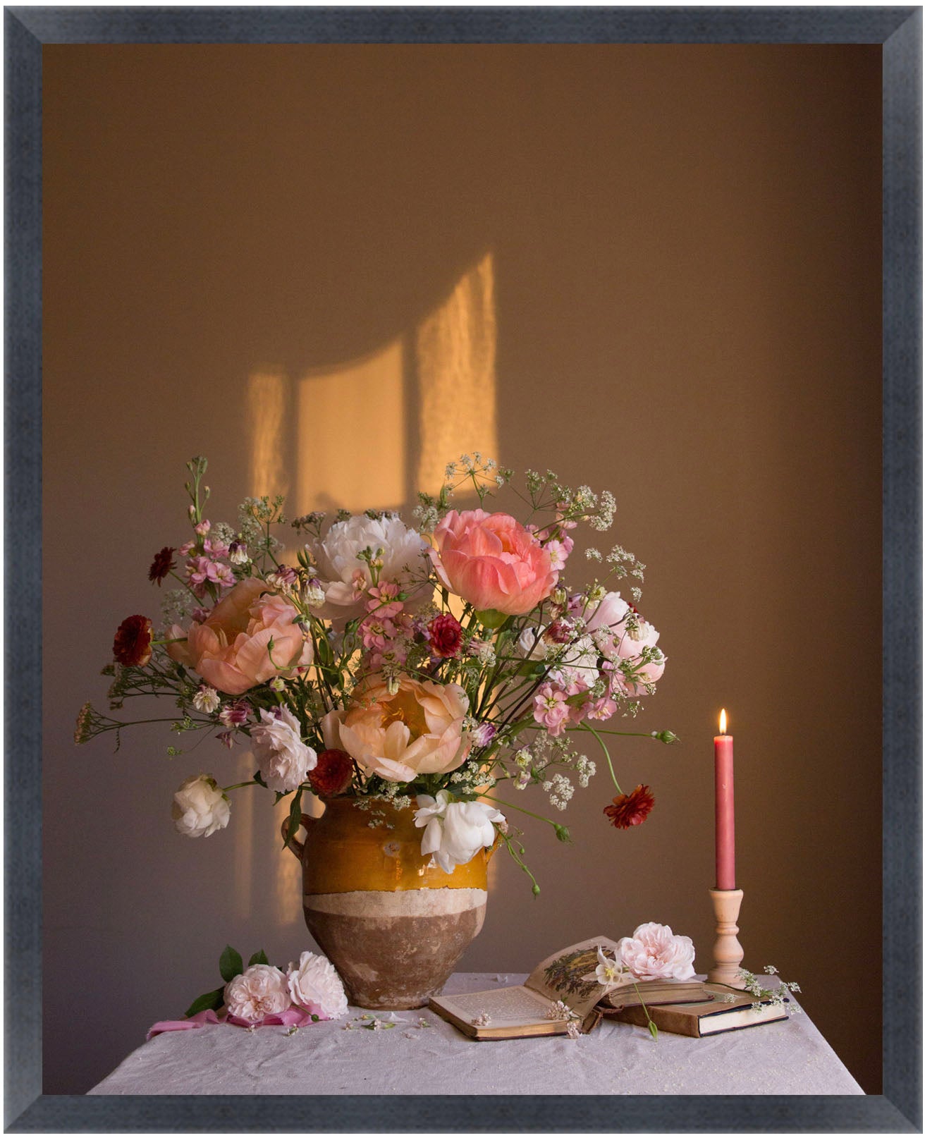 Floral arrangement in a vase with a candle and books on a table against a brown wall.   Flower still-life