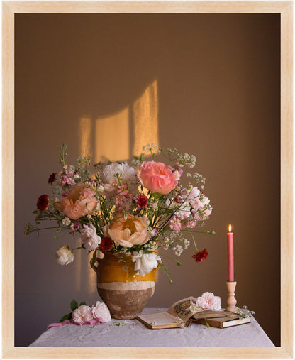 Floral arrangement in a vase with a candle and books on a table against a brown wall.  Flower print