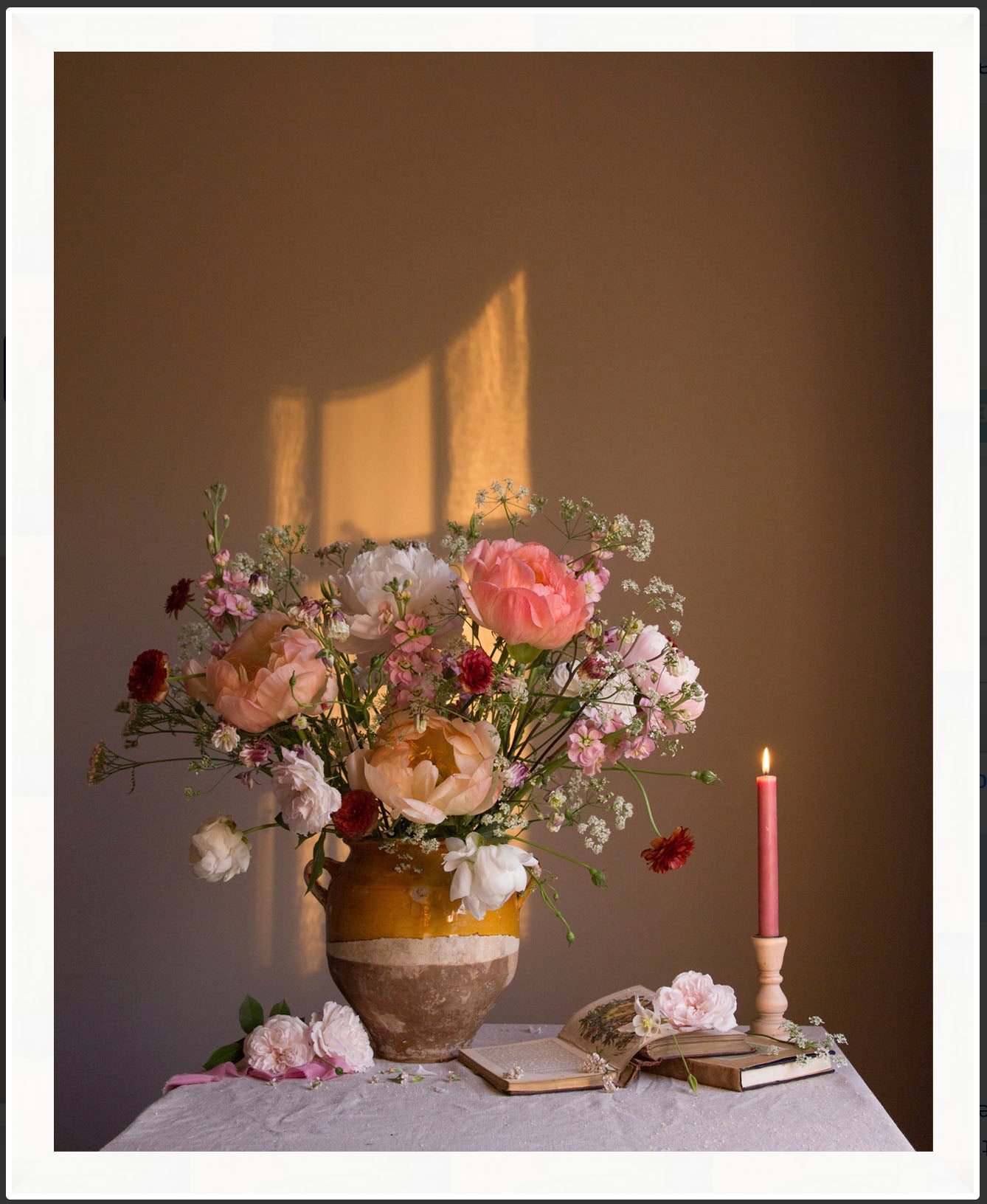 Floral Still-life with a candle and books on a table against a brown wall.