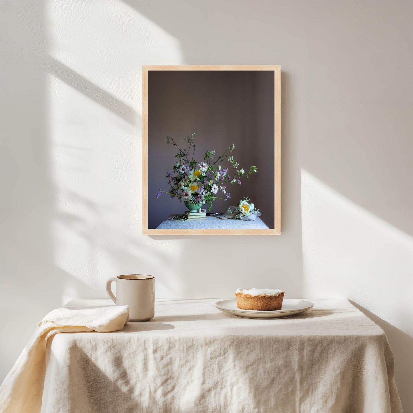 Framed artwork of a floral arrangement on a wall above a table with a pie and mug.   Floral artwork