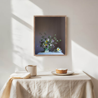 Framed artwork of a floral arrangement on a wall above a table with a pie and cup.   Botanical artwork