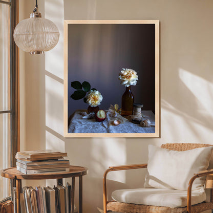 Framed artwork of a still life scene with flowers and a bottle on a wall in a room with a chair and books.   Still life photograph