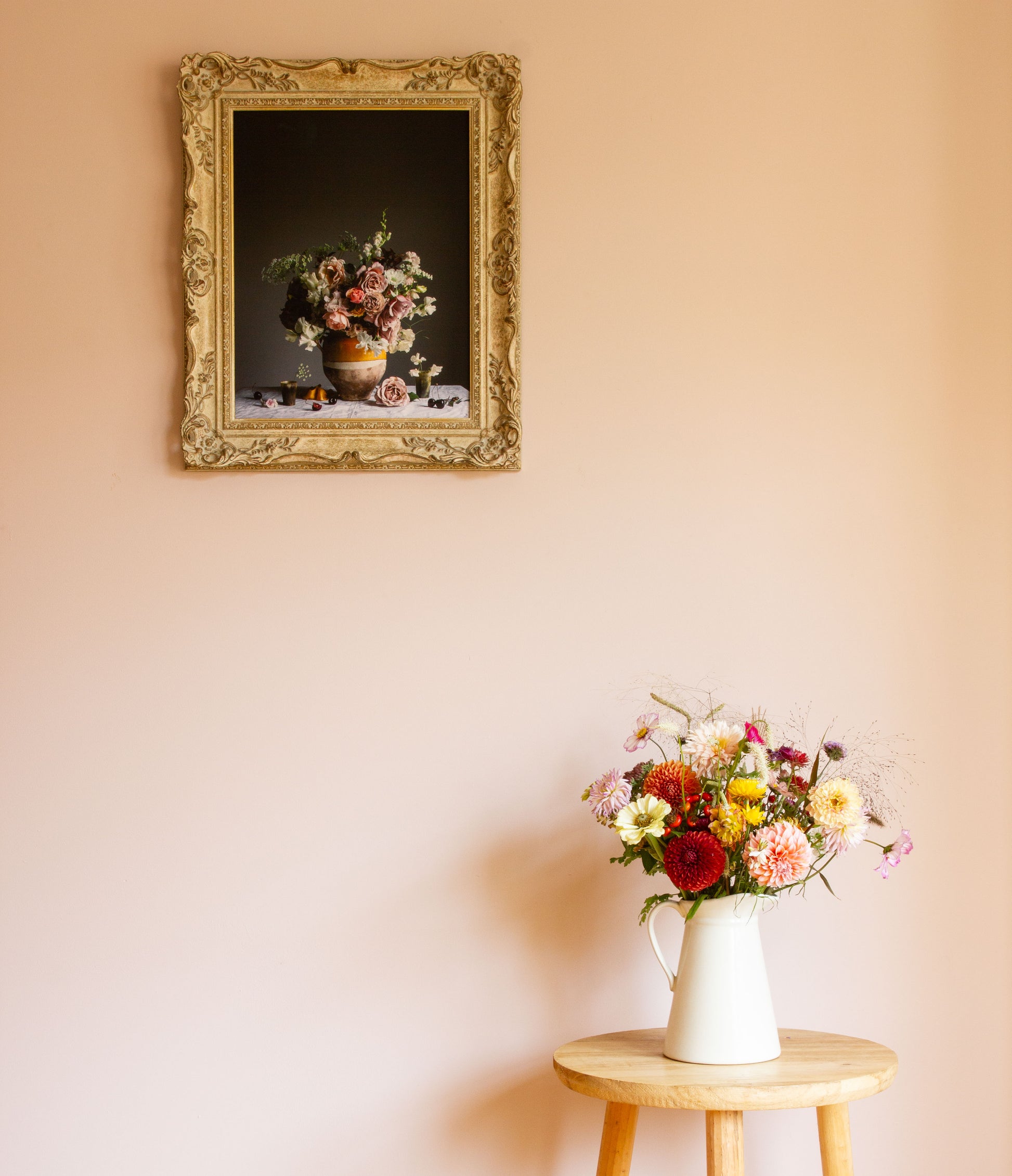 Floral arrangement in a white vase on a small wooden table with a framed limited edition still-life of flowers on the wall.